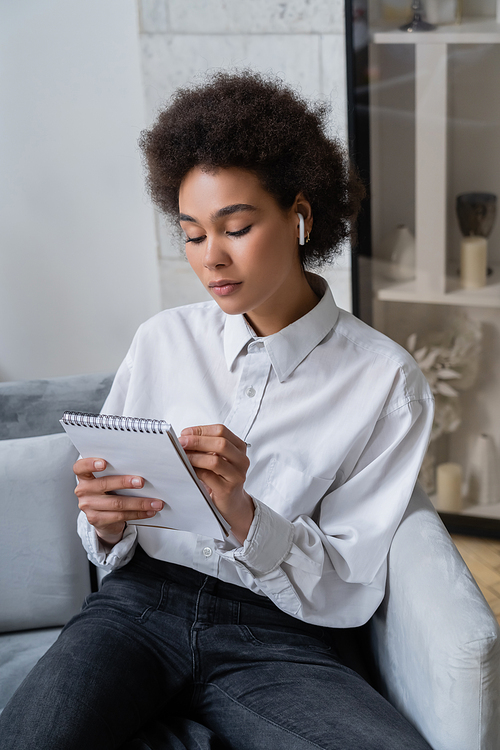 curly african american woman in white shirt and wireless earphone making notes in notebook