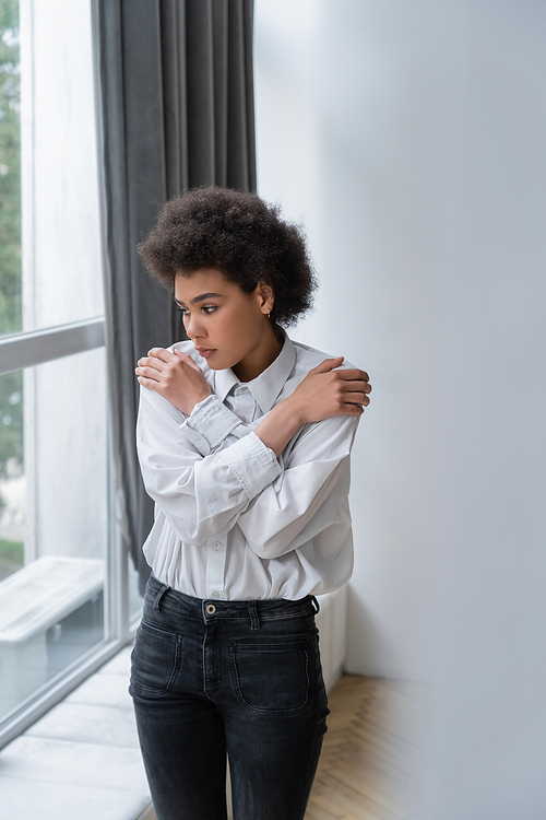 depressed african american woman in white shirt and jeans hugging shoulders at home
