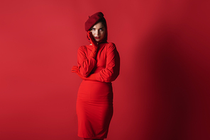 young elegant woman in beret and dress posing while looking at camera on red