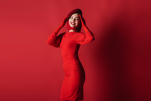 joyful and elegant woman in beret and dress posing on red
