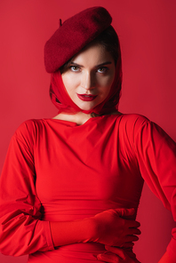 elegant young woman in beret and dress looking at camera isolated on red
