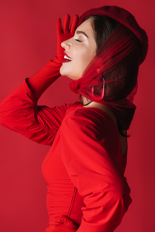 side view of happy young woman in beret and dress isolated on red