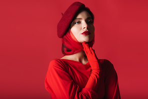 elegant young woman in beret and glove looking away and posing isolated on red
