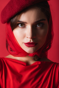 close up of elegant young woman in beret and headscarf looking at camera isolated on red
