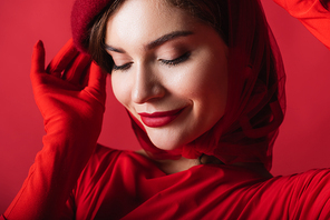 close up of smiling young woman adjusting beret isolated on red