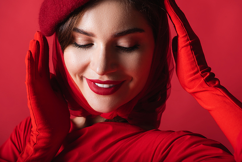 cheerful young woman adjusting beret isolated on red