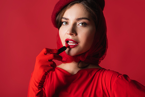 young woman in glove, headscarf and beret applying lipstick isolated on red