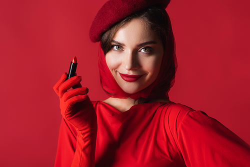 happy woman in glove, headscarf and beret holding lipstick isolated on red