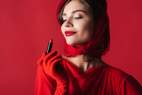 cheerful woman in glove, headscarf and beret looking at lipstick isolated on red