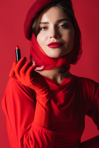 young woman in headscarf and beret holding lipstick isolated on red