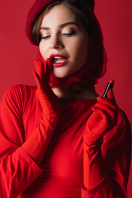 pretty woman in gloves, headscarf and beret holding lipstick isolated on red