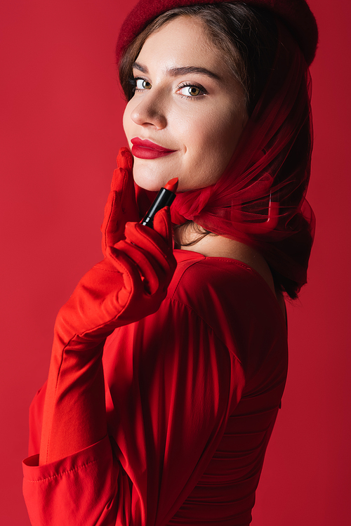 pleased young woman in gloves, headscarf and beret holding lipstick isolated on red