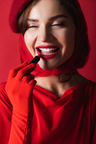 joyful woman in glove, headscarf and beret applying lipstick isolated on red