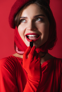 excited woman in glove, headscarf and beret applying lipstick isolated on red