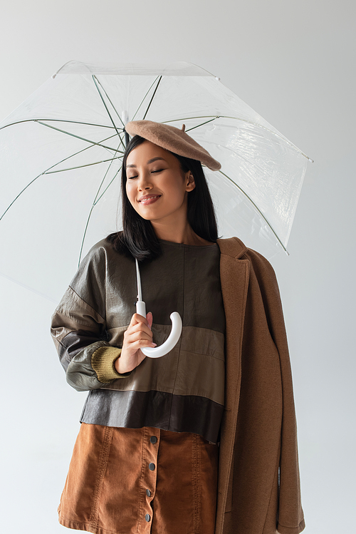 trendy asian woman standing under transparent umbrella and smiling with closed eyes isolated on grey