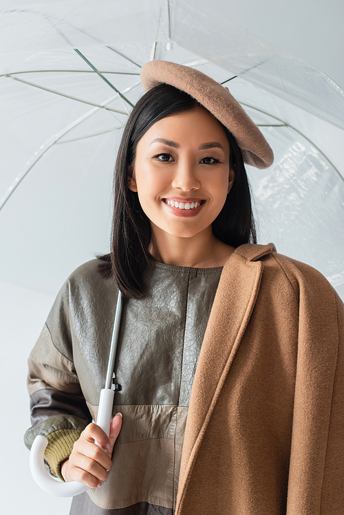 happy asian woman in beret looking at camera under transparent umbrella isolated on grey