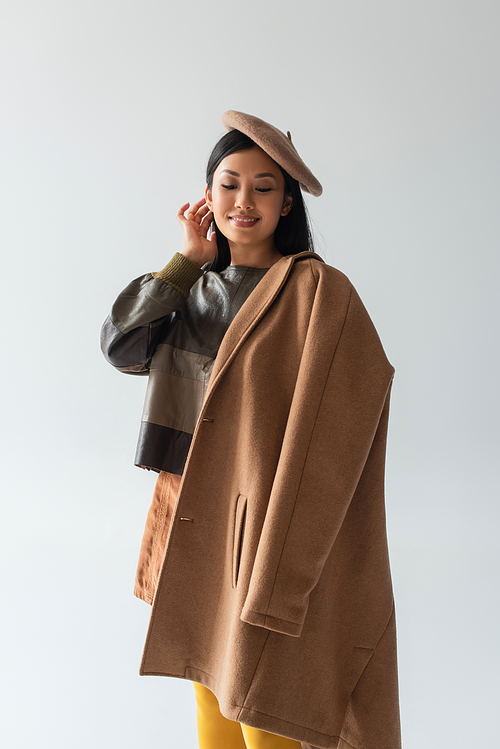 joyful asian woman with beige coat touching hair isolated on grey