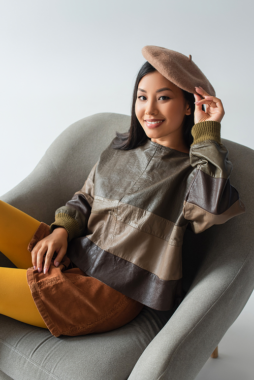 asian woman in leather jacket and beret sitting in armchair on grey