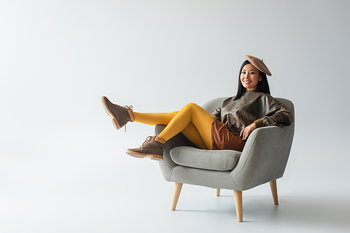 full length view of happy asian woman in leather pullover and yellow tights sitting in armchair on grey