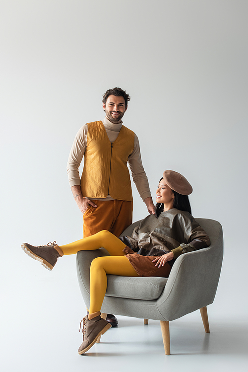 happy and stylish man standing with hand in pocket near asian woman in armchair on grey