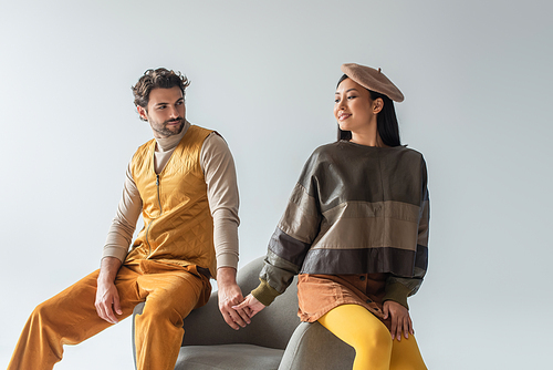 smiling asian woman in beret and leather pullover holding hands with man in yellow vest isolated on grey