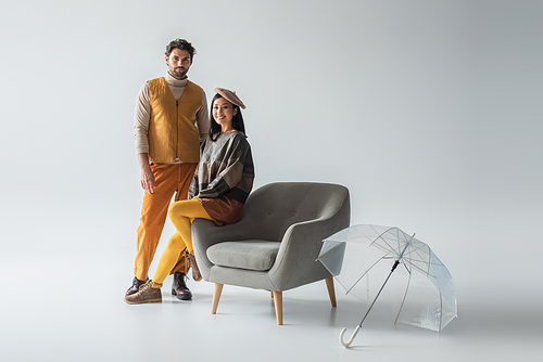 cheerful asian woman sitting on armchair near trendy man and transparent umbrella on grey