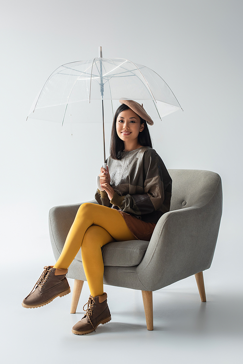 smiling asian woman in leather pullover and yellow tights sitting in armchair under transparent umbrella on grey