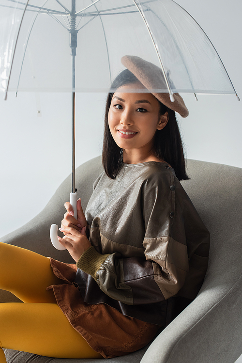 stylish asian woman sitting in armchair under umbrella and smiling at camera isolated on grey