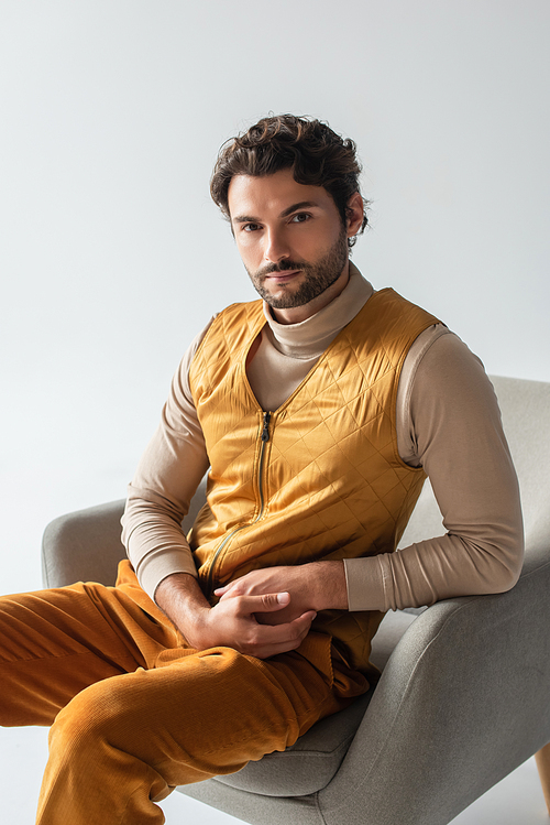 stylish man in yellow vest and trousers sitting in armchair and looking at camera isolated on grey