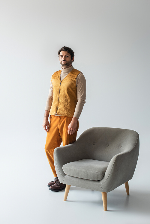 full length view of man in yellow trousers and vest standing near armchair on grey