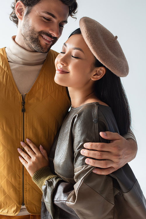 happy and stylish man embracing asian woman smiling with closed eyes isolated on grey