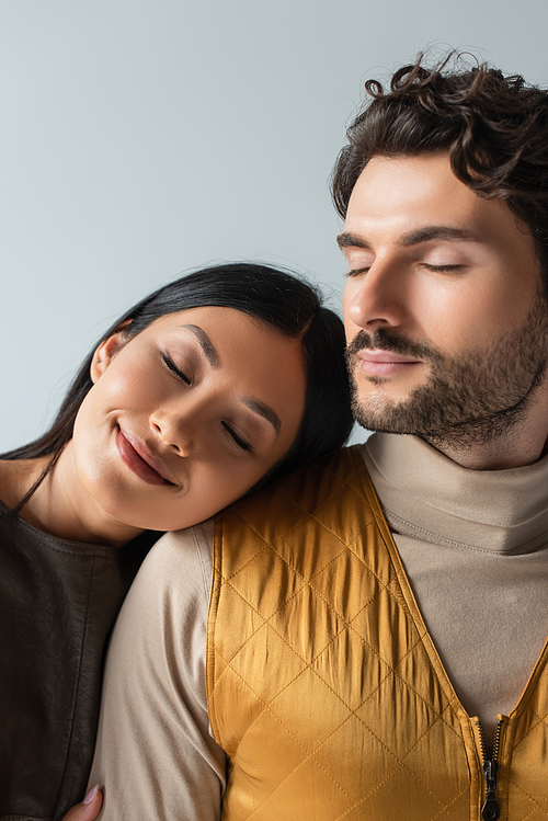 smiling asian woman with closed eyes leaning on stylish brunette man isolated on grey