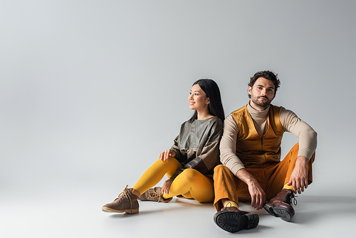 trendy man smiling at camera while sitting near happy asian woman on grey