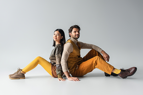 stylish interracial couple smiling at camera while sitting back to back on grey