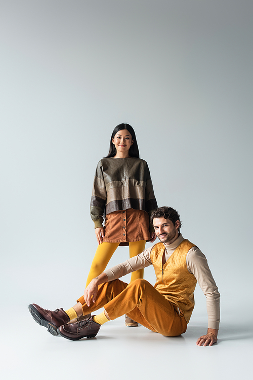 smiling man in yellow trousers sitting near asian woman in leather jacket on grey