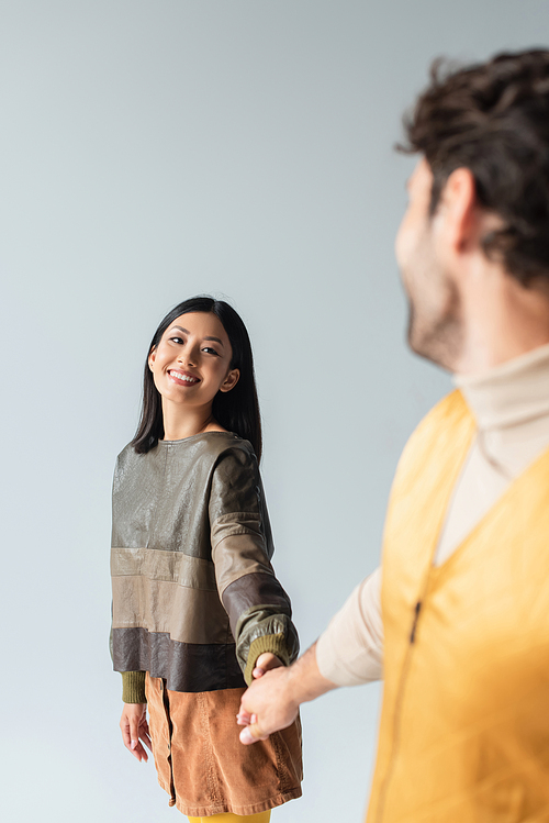 cheerful asian woman in leather pullover holding hands with blurred man isolated on grey