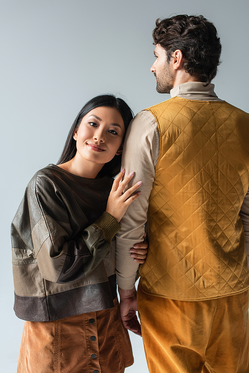 happy asian woman in leather pullover leaning on man in yellow vest isolated on grey