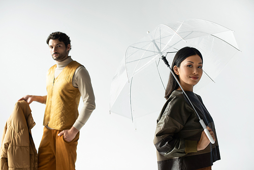 asian woman smiling at camera under transparent umbrella near man holding puffer jacket on grey