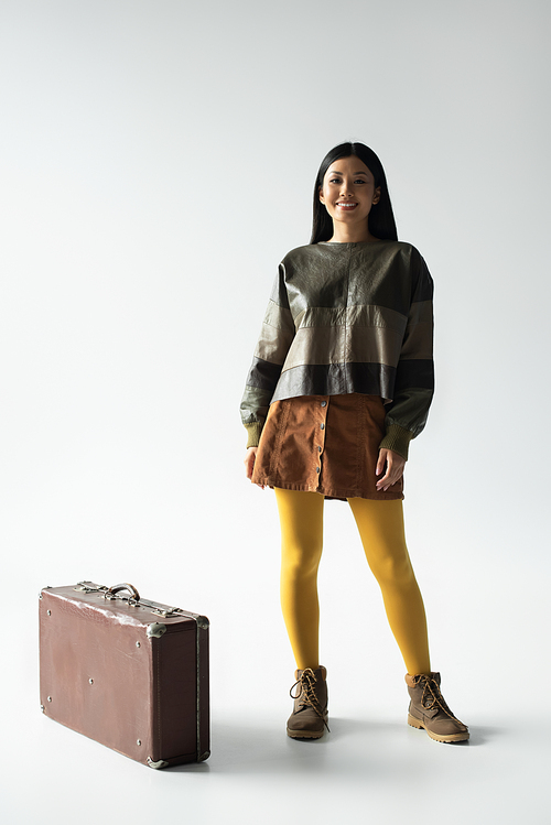 happy asian woman in stylish outfit posing near vintage suitcase on grey