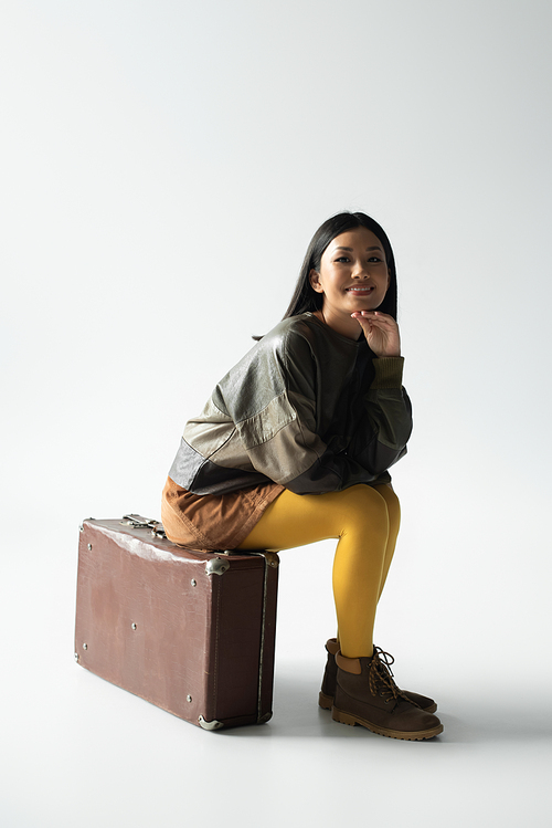 asian woman in trendy clothing sitting on vintage suitcase on grey