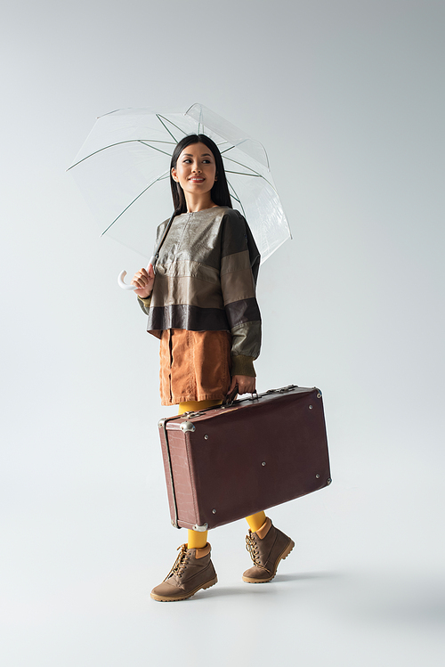 full length view of stylish asian woman with vintage suitcase walking under transparent umbrella on grey