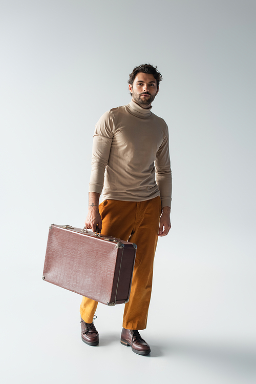 full length view of man in beige turtleneck and yellow trousers walking with vintage suitcase on grey