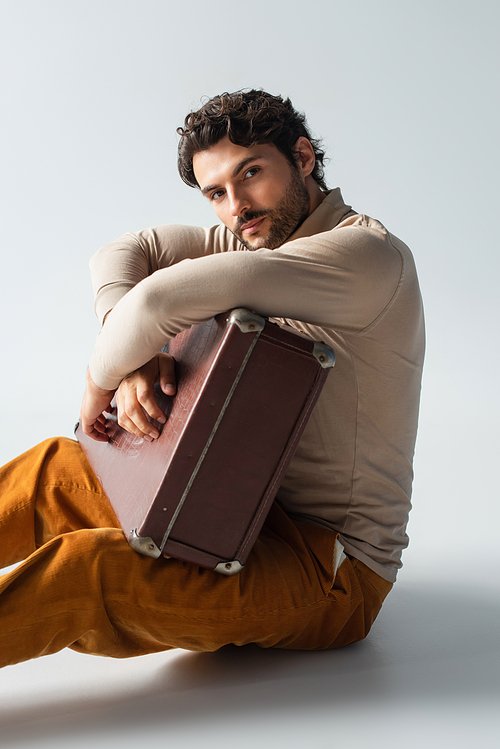 trendy man in yellow trousers and beige turtleneck sitting with vintage suitcase on grey