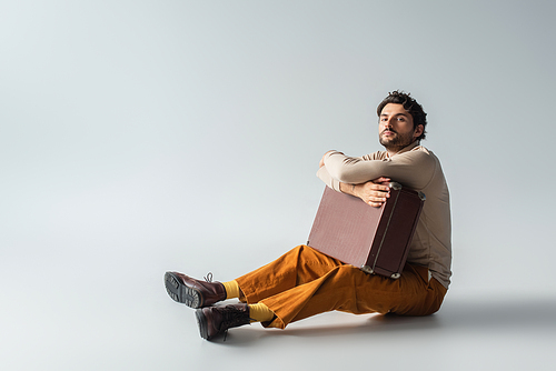 full length view of man in trendy clothes sitting with vintage suitcase on grey