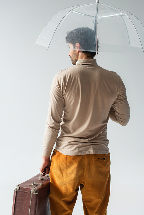 back view of stylish man with vintage suitcase standing under transparent umbrella isolated on grey