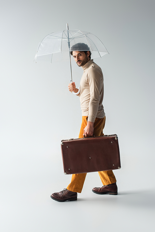 fashionable man with vintage suitcase walking under transparent umbrella on grey