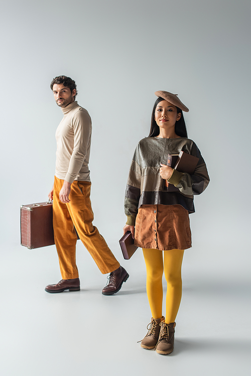 full length view of man with vintage suitcase and asian woman with books on grey