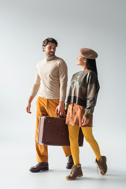 trendy interracial couple carrying vintage suitcase and smiling at each other on grey