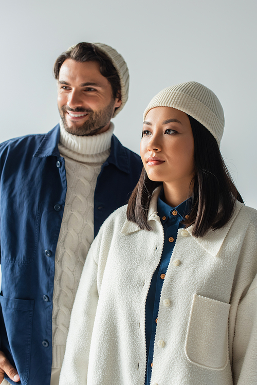 asian woman in white jacket and beanie near trendy man smiling on blurred background isolated on grey