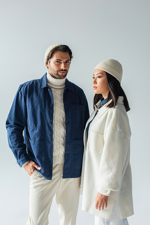 man in blue jacket standing with hand in pocket near trendy asian woman isolated on grey